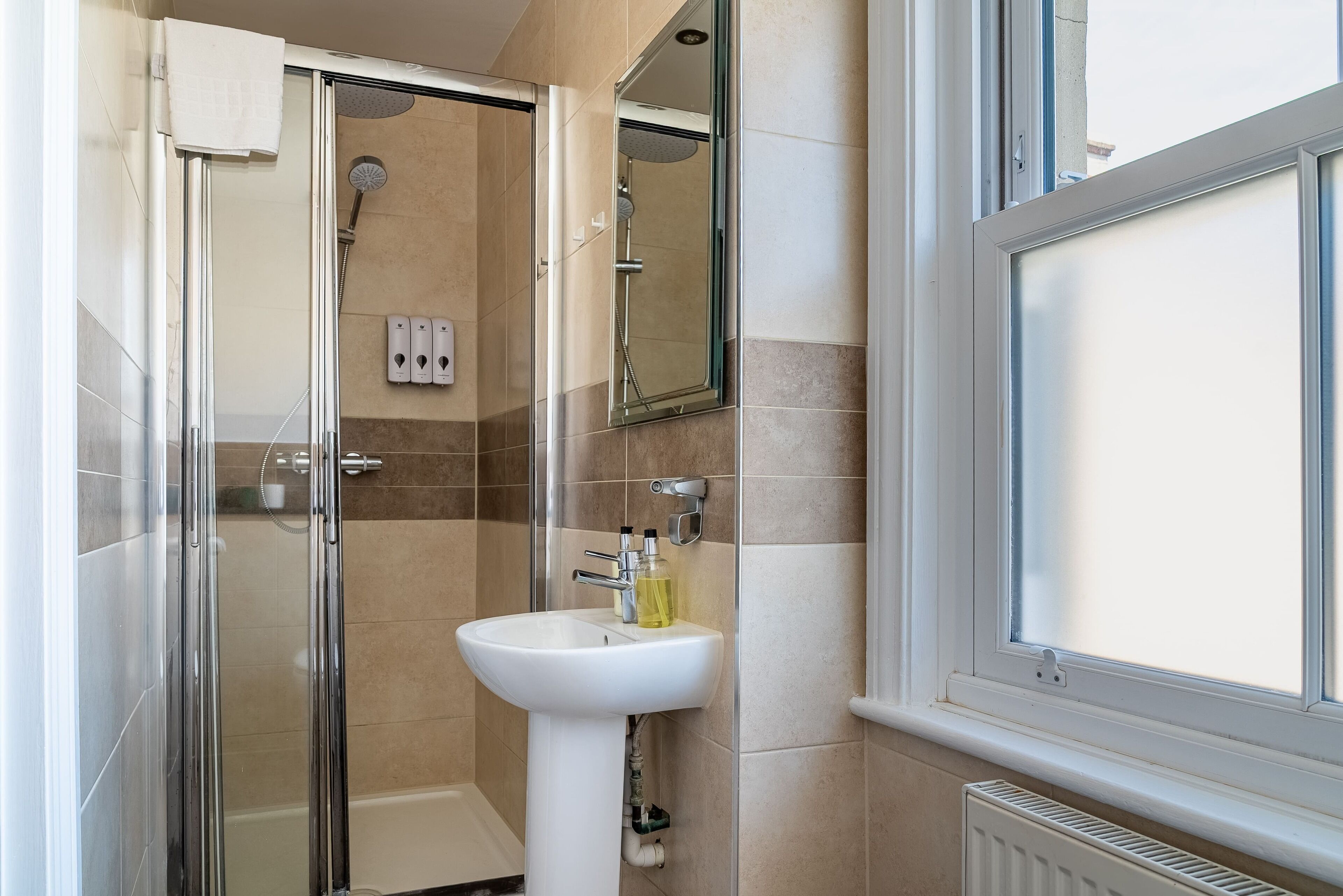 A modern bathroom with a glass shower enclosure, a white pedestal sink, and a window.