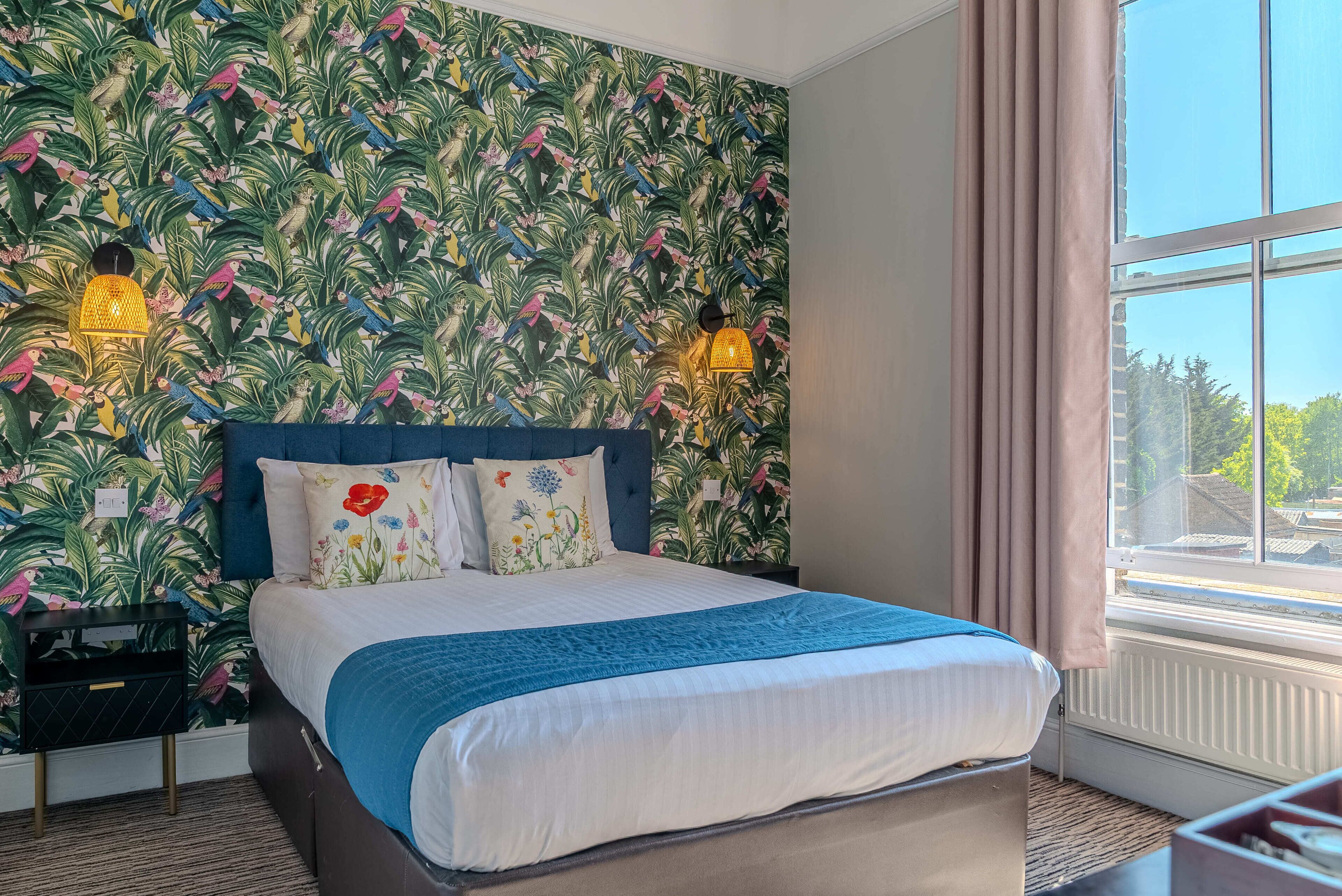 Un dormitorio con cama, papel tapiz floral, dos lámparas fijadas a la pared, una ventana con cortinas y vistas a árboles fuera.