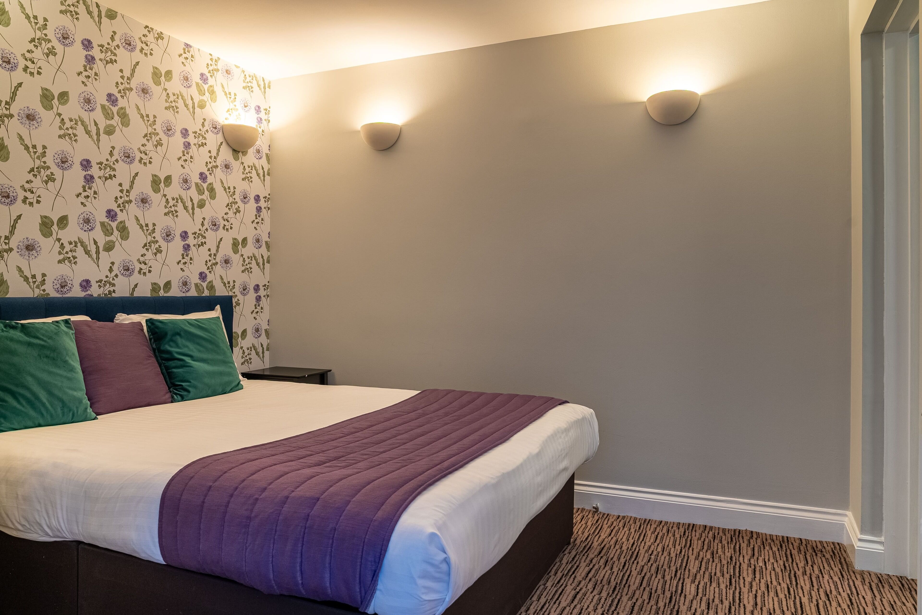 A neatly made bed with a purple blanket, a floral wallpaper, and two wall-mounted lights.