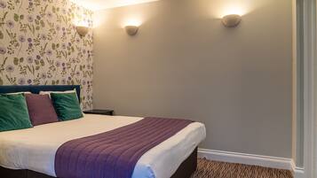 A neatly made bed with a purple blanket, a floral wallpaper, and two wall-mounted lights.