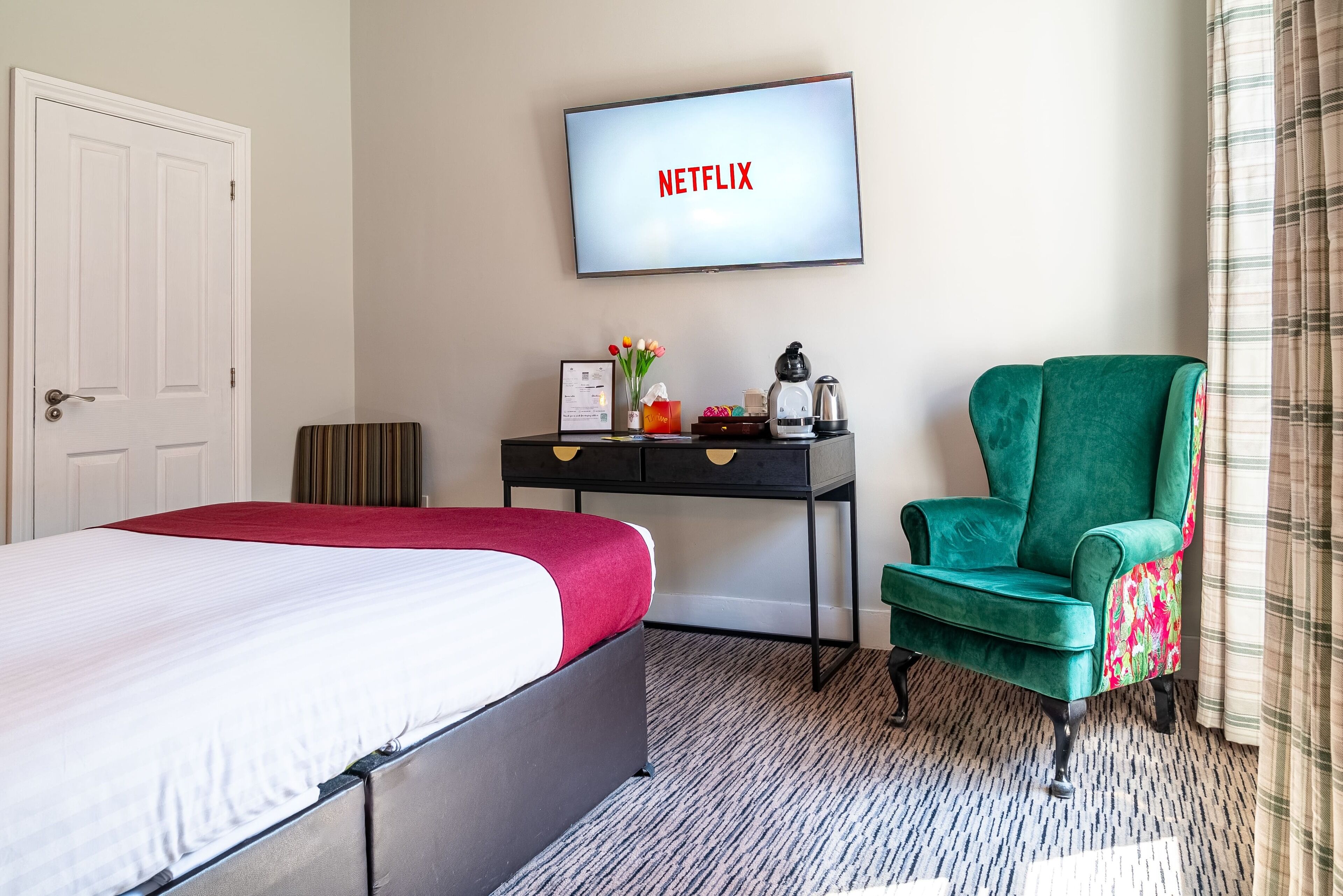 Habitación de hotel con cama, escritorio con computadora, sillón verde y televisor mostrando 'NETFLIX'.