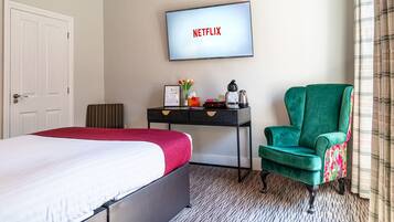 Habitación de hotel con cama, escritorio con computadora, sillón verde y televisor mostrando 'NETFLIX'.