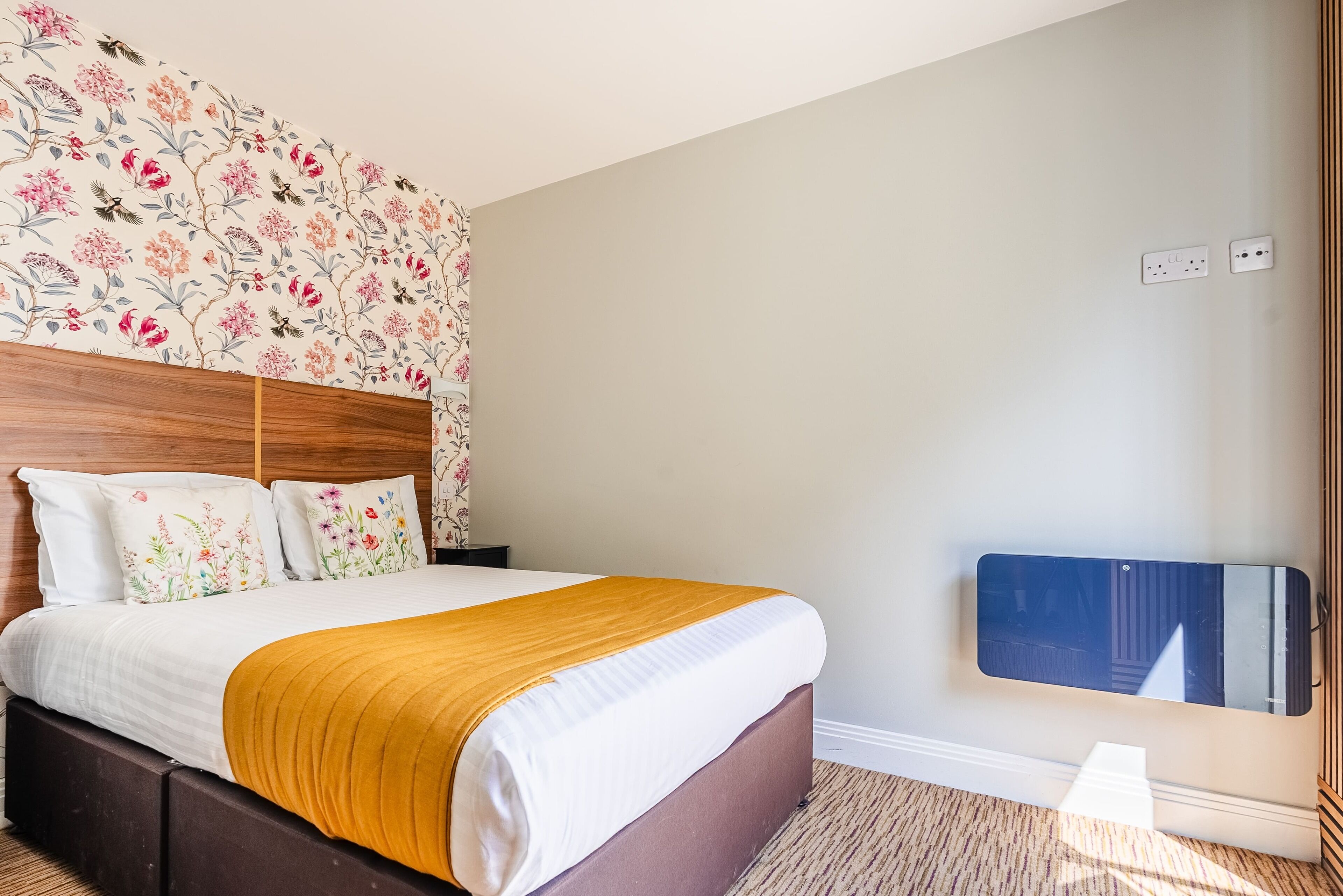 A neatly made bed with a wooden headboard, a floral-patterned wall, and a mirror on the wall.