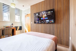 Une chambre d’hôtel dotée d’un grand téléviseur à écran plat fixé au mur, d’un lit avec une couvre-lit à rayures, d’un bureau en bois avec une chaise et d’une fenêtre avec des stores.