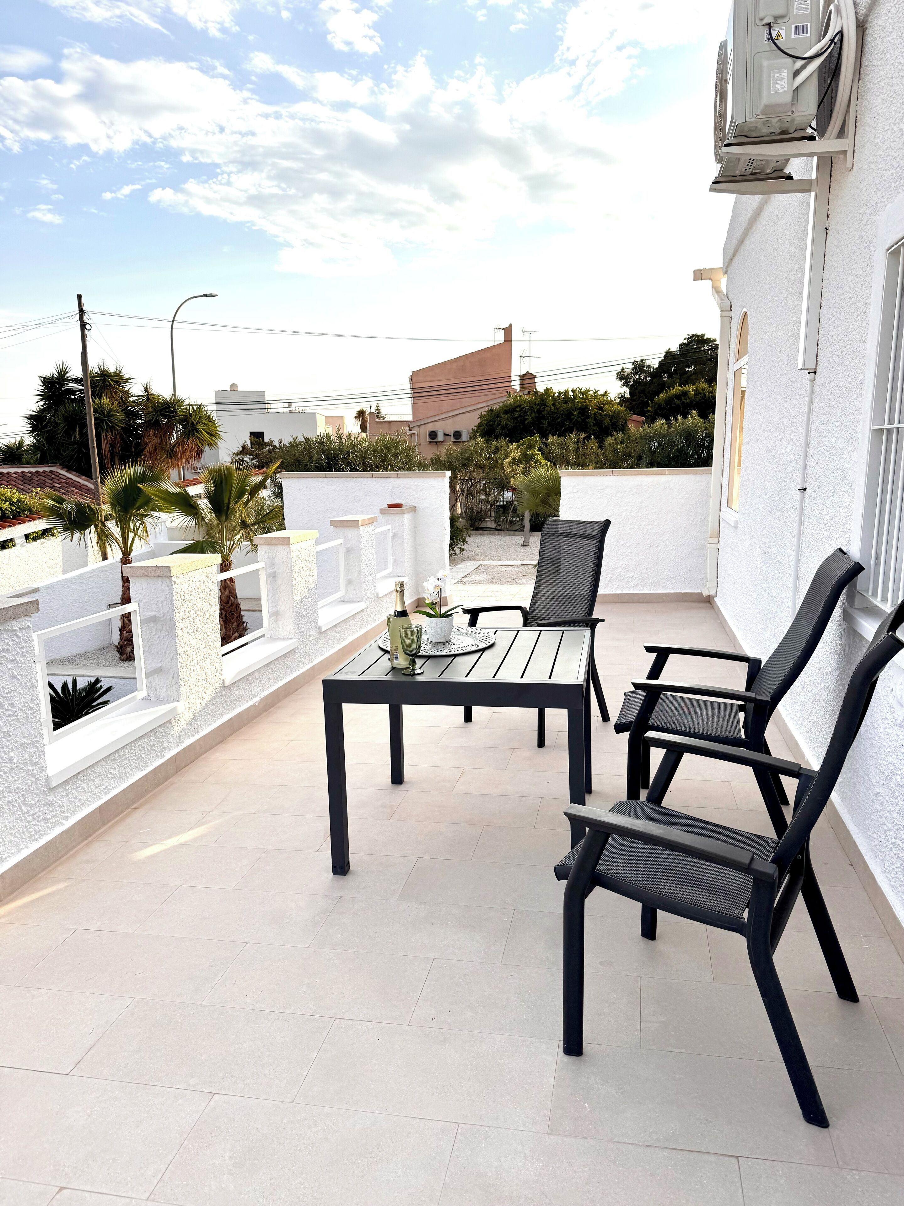 An outdoor patio with a table and chairs, a white wall, and a view of buildings and trees.