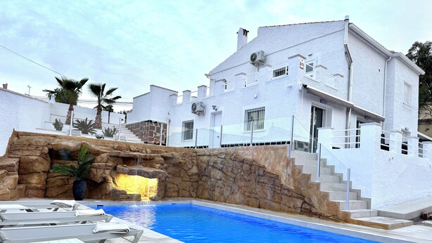 A swimming pool with a rock feature, lounge chairs, and a white building with a stone wall.