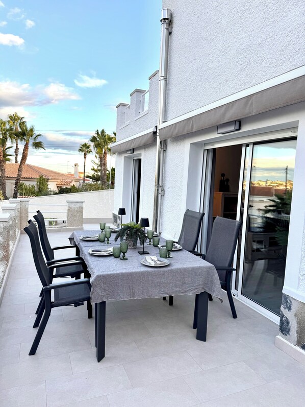 An outdoor dining area with a table set for four, surrounded by chairs, on a terrace with a view of palm trees and a building in the background. - Luxurious detached villa, private pool in a privileged location (San Fulgencio)