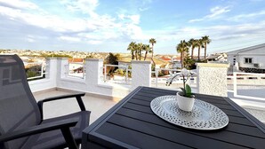 A balcony with a table and chair, overlooking a coastal landscape with palm trees and buildings.