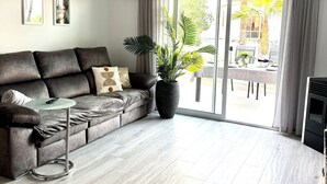 A modern living room with a grey sofa, a glass coffee table, a large potted plant, and a sliding glass door leading to an outdoor area.