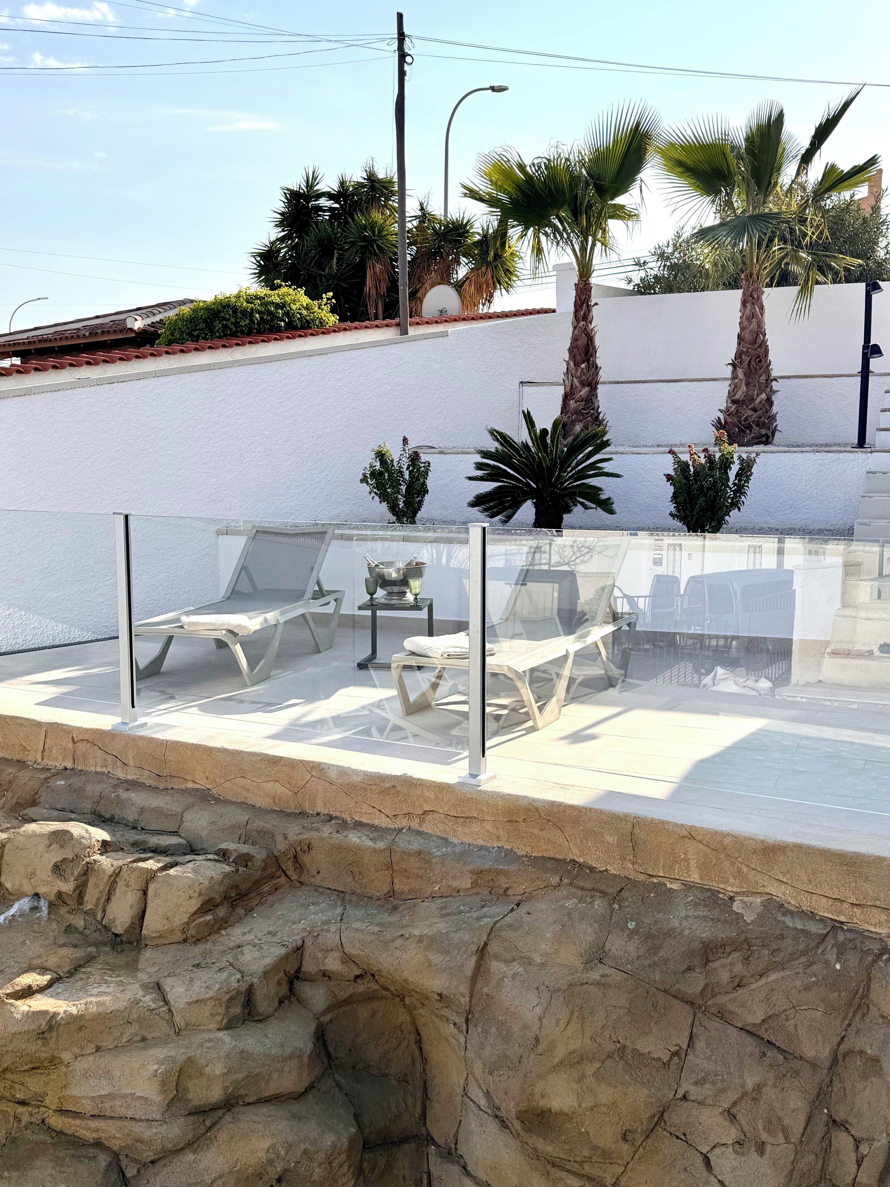 Une terrasse sur le toit, dotée de chaises longues et d’une table, entourée de palmiers et d’un mur blanc.