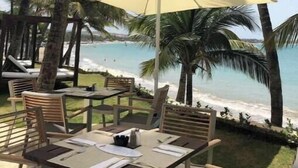 Een terras aan het strand met een tafel, stoelen, een grote parasol en palmbomen.
