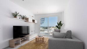 A modern living room with a flat-screen TV, a grey sofa, a wooden coffee table, and a large window offering a view of the city. - Hacienda Casares 2053 Geranio (Casares)