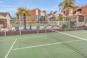 A tennis court with a net, surrounded by a fence, and a pool area with lounge chairs.
