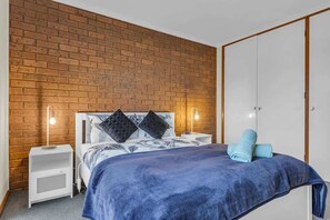 Une chambre avec un mur en briques, un lit recouvert d’une couverture bleue, deux tables de chevet avec des lampes et une armoire.