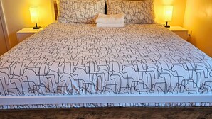 A bedroom with a large bed, two bedside tables with lamps, a wooden floor, and a painting on the wall. - Spacious & Convenient 1BR unit-20min to Boston (Melrose)