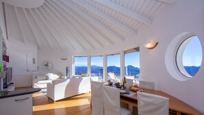 A spacious room with a wooden floor, white furniture, a dining table, and large windows offering a mountain view. - The Perfect View Loft (Premeno)