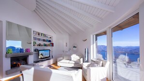 A modern living room with a fireplace, white furniture, a bookshelf, and a large window with a mountain view. - The Perfect View Loft (Premeno)