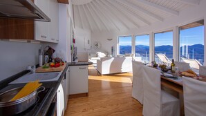 A modern kitchen with wooden cabinets, a dining area with white furniture, and a view of the mountains through large windows. - The Perfect View Loft (Premeno)