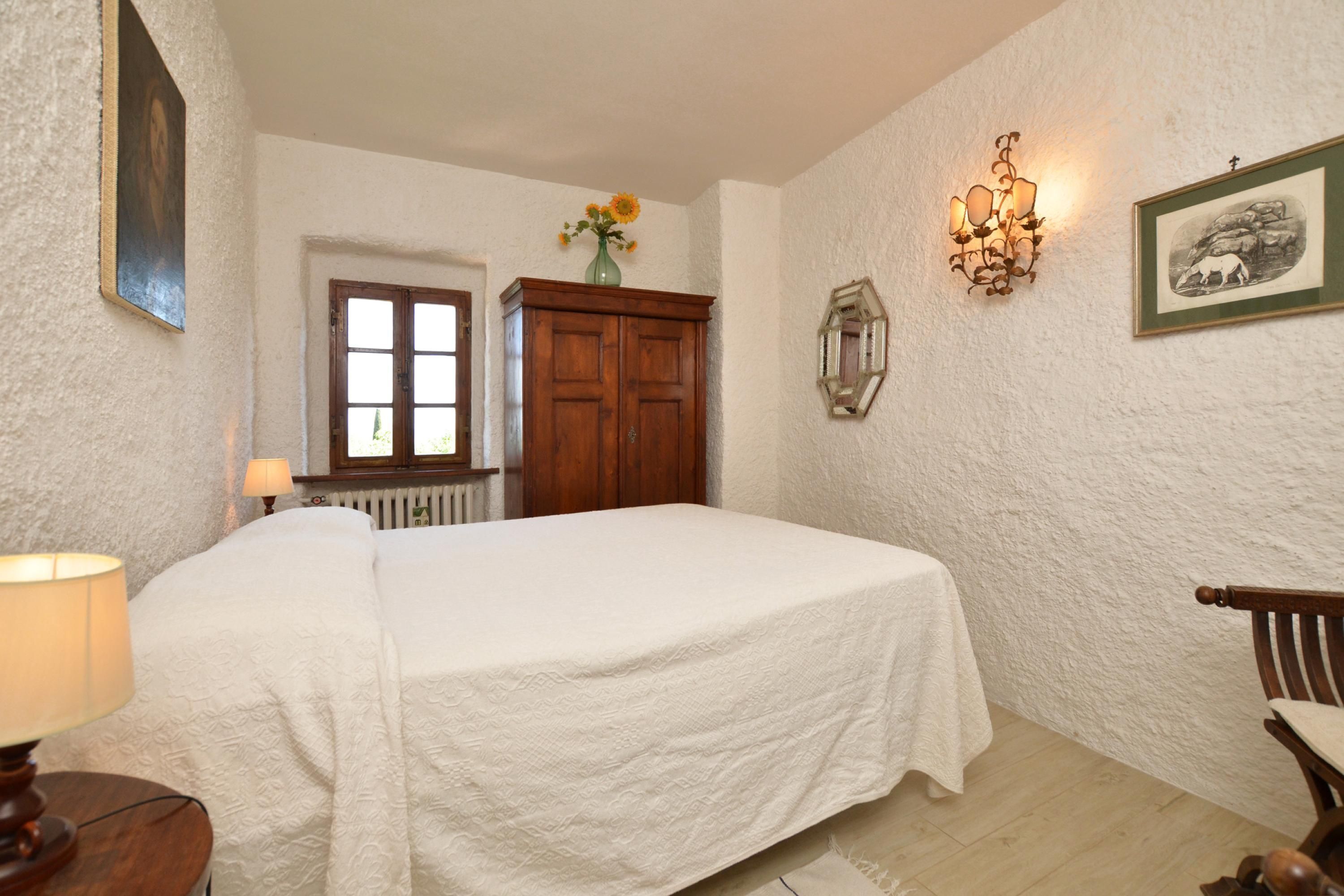 A bedroom with a bed, a wooden wardrobe, a nightstand, a lamp, a vase with flowers, a wall-mounted light fixture, and a framed picture.