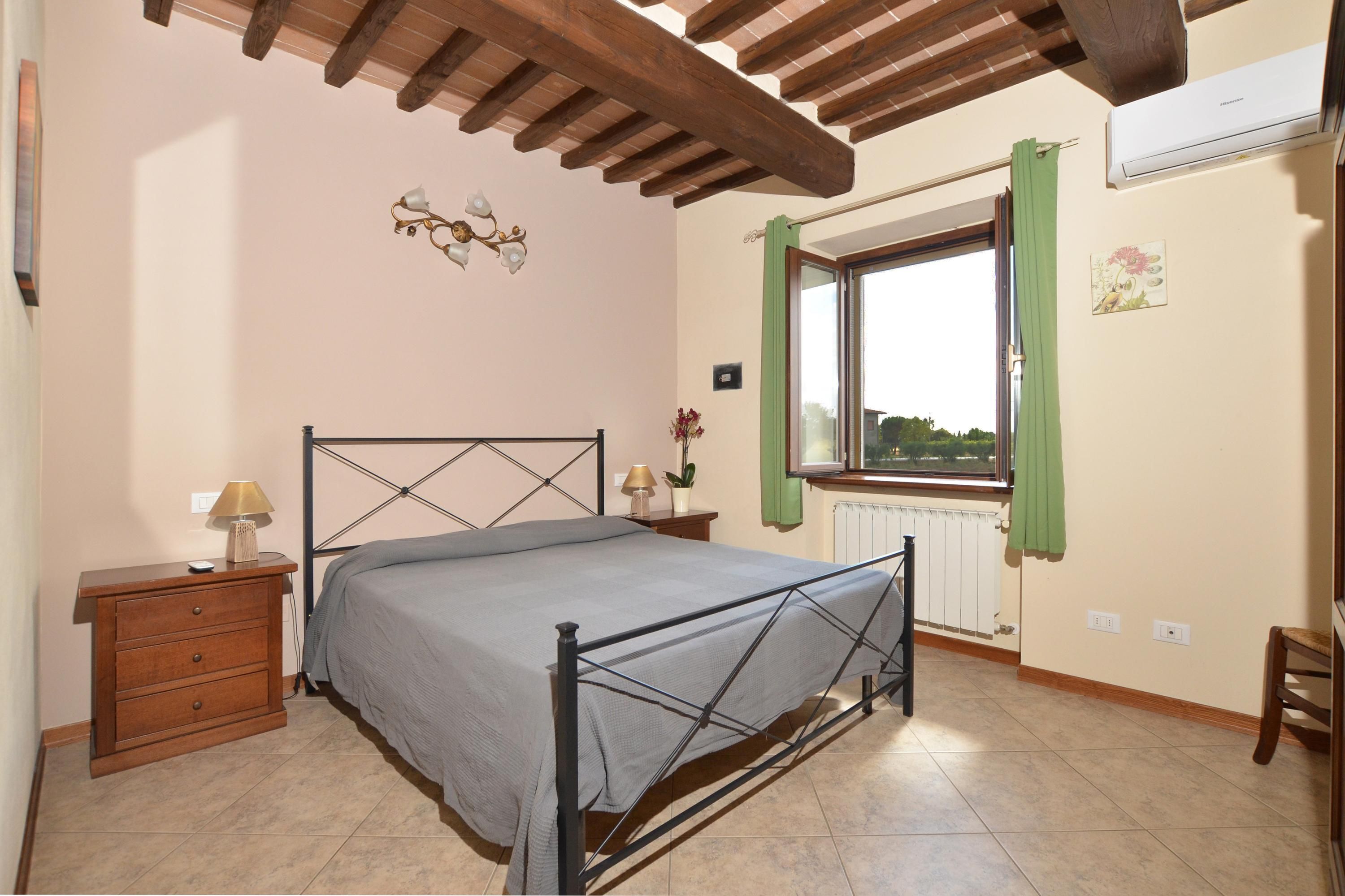 A bedroom with a bed, nightstand, lamp, window with curtains, radiator, and a wall-mounted air conditioner.