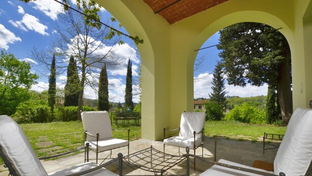 A covered outdoor seating area with white cushioned chairs and a metal table, surrounded by greenery and trees.