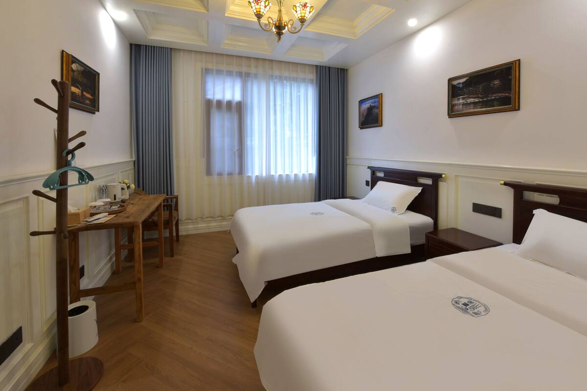 A hotel room with two beds, a wooden desk, a chair, a coat rack, and a chandelier.