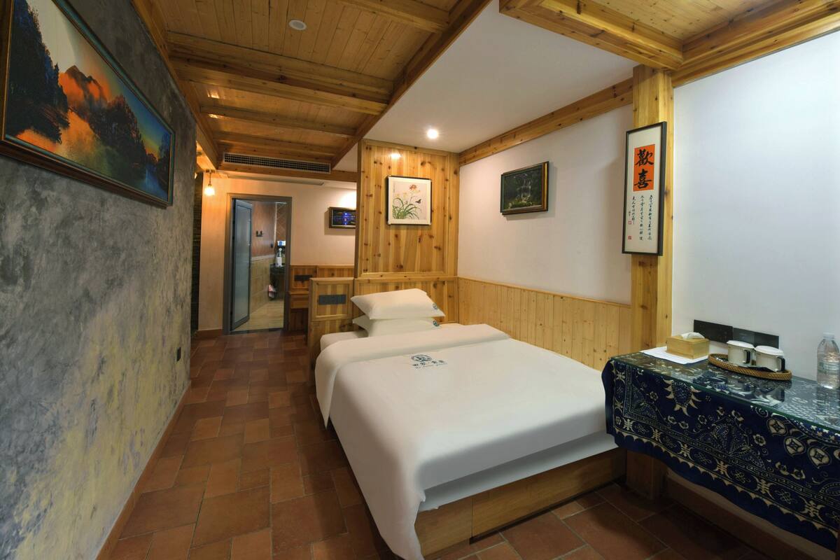 A hotel room with a wooden ceiling, a bed with white bedding, a wooden headboard, a small table with a patterned tablecloth, and a wall with framed artwork.