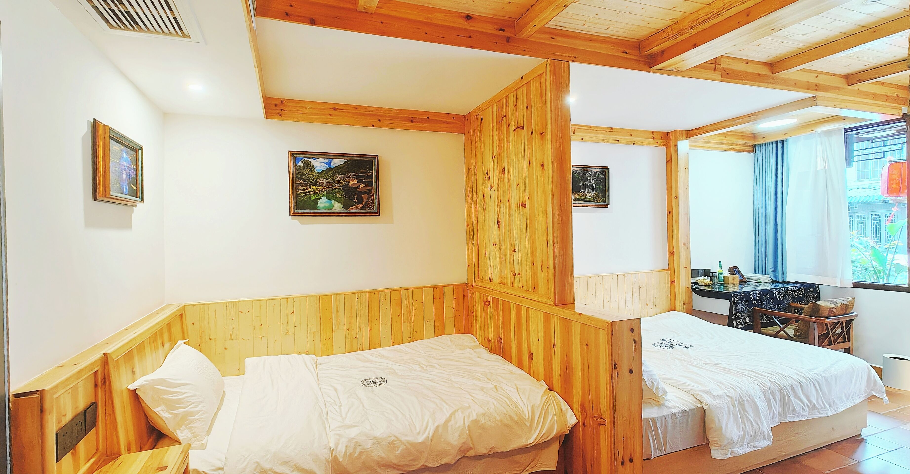 A room with wooden paneling, two beds, a small table with a chair, and framed pictures on the walls.