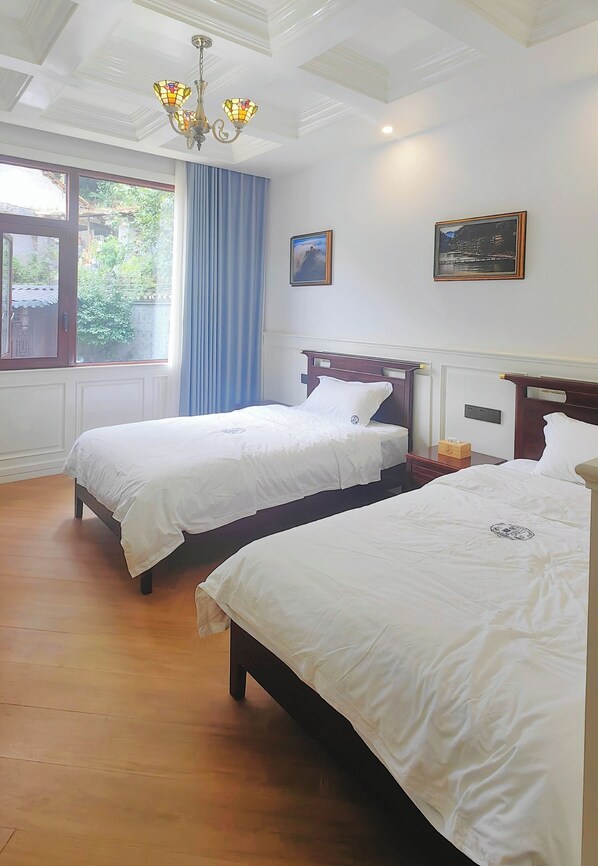 A hotel room with two beds, a chandelier, a window with curtains, and framed pictures on the wall.