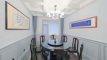 Uma sala de jantar formal com uma mesa redonda de vidro, cadeiras de madeira, um candelabro e obras de arte emolduradas nas paredes.