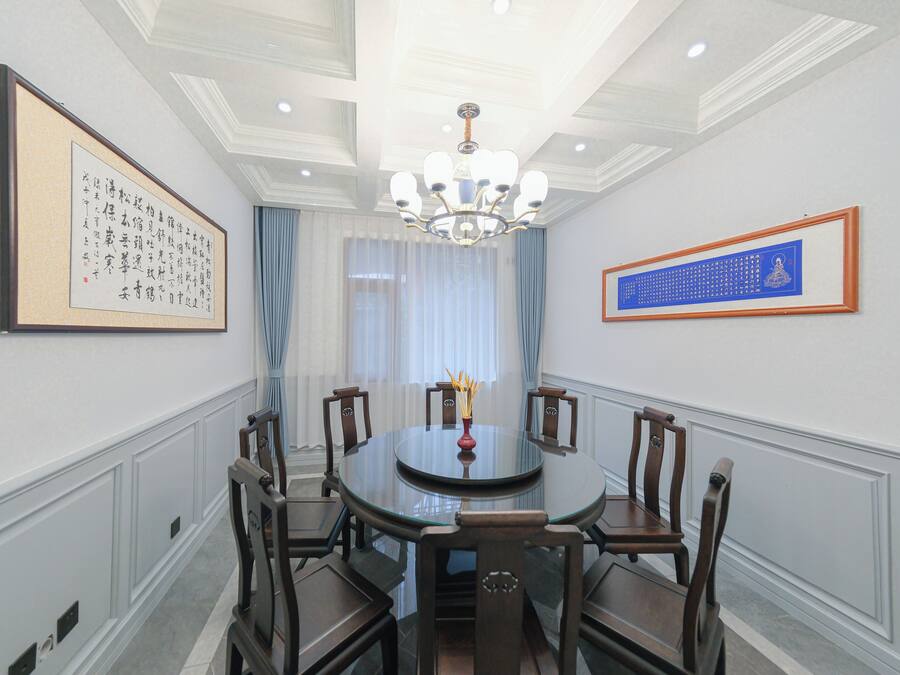 Uma sala de jantar formal com uma mesa redonda de vidro, cadeiras de madeira, um candelabro e obras de arte emolduradas nas paredes.