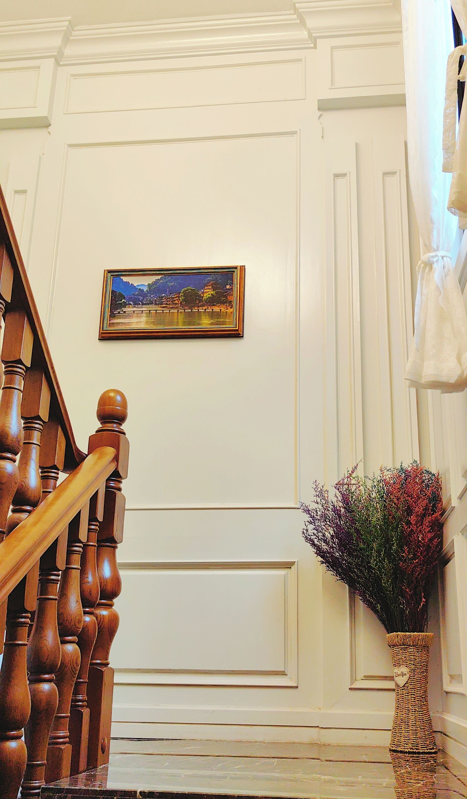 A staircase with wooden balusters, a framed landscape painting on the wall, and a vase with dried flowers.