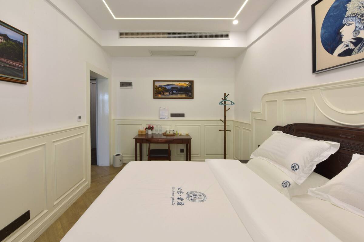A hotel room with a large bed, a desk with a chair, a painting on the wall, and a coat rack.