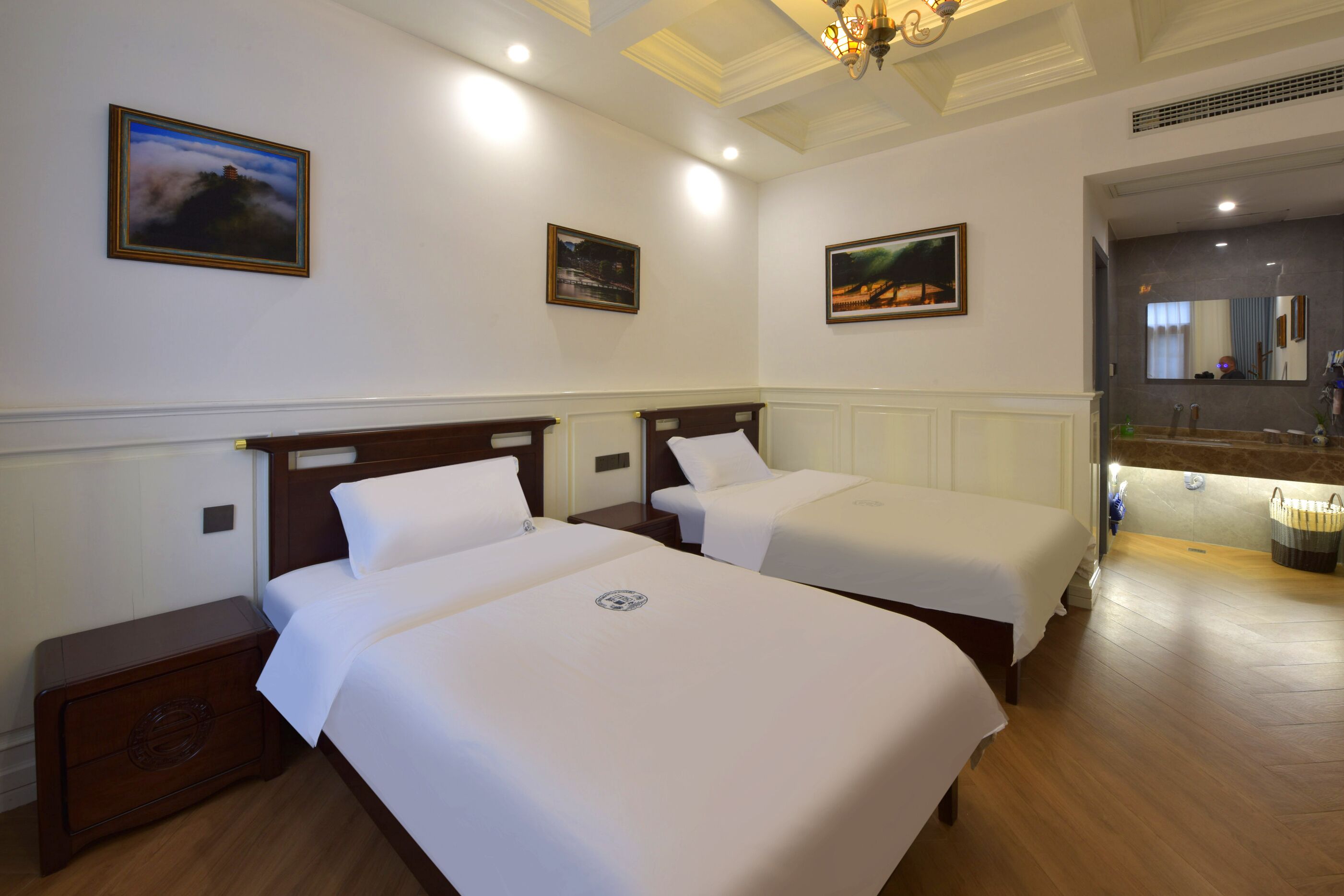A hotel room with two single beds, a wooden nightstand, a bathroom with a sink and mirror, and framed pictures on the walls.