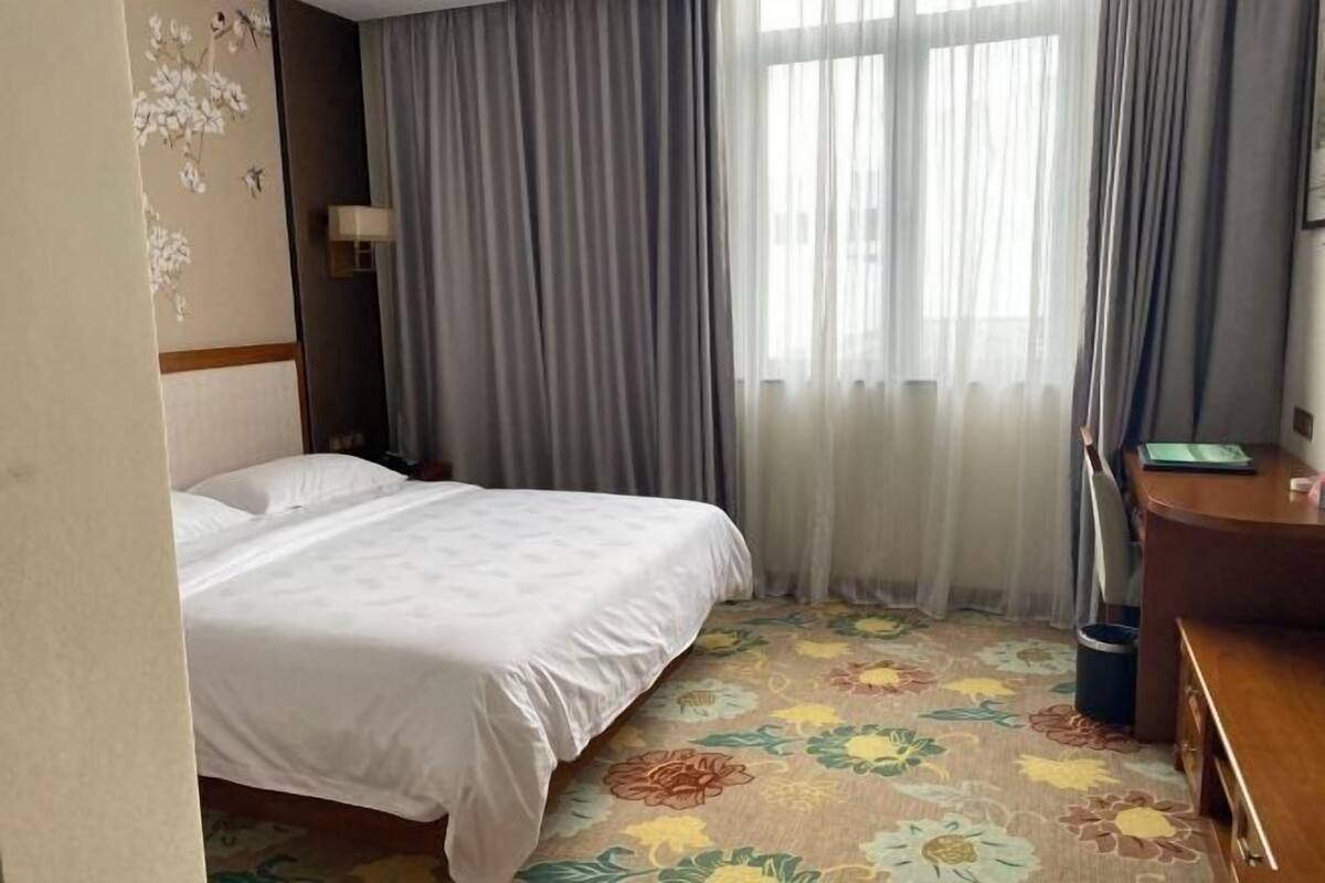 Habitació d'hotel amb llit, escriptori, cadira, làmpada i finestra amb cortines.