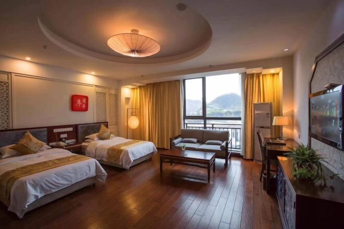Habitació d'hotel amb dos llits, un sofà, una taula de cafè, televisió i una finestra gran amb cortines.