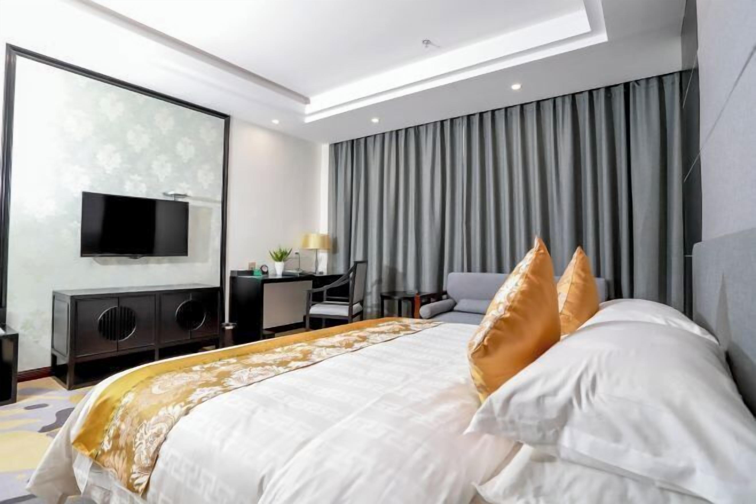 A modern hotel room with a large bed, a flat-screen TV mounted on the wall, a desk with a chair, and a large window with curtains.