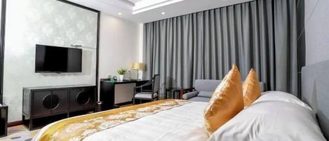 A modern hotel room with a large bed, a flat-screen TV mounted on the wall, a desk with a chair, and a large window with curtains.