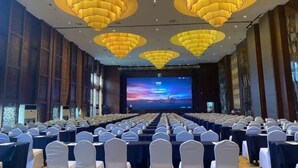 A conference room with rows of tables and chairs, a large screen at the front, and decorative ceiling lights.