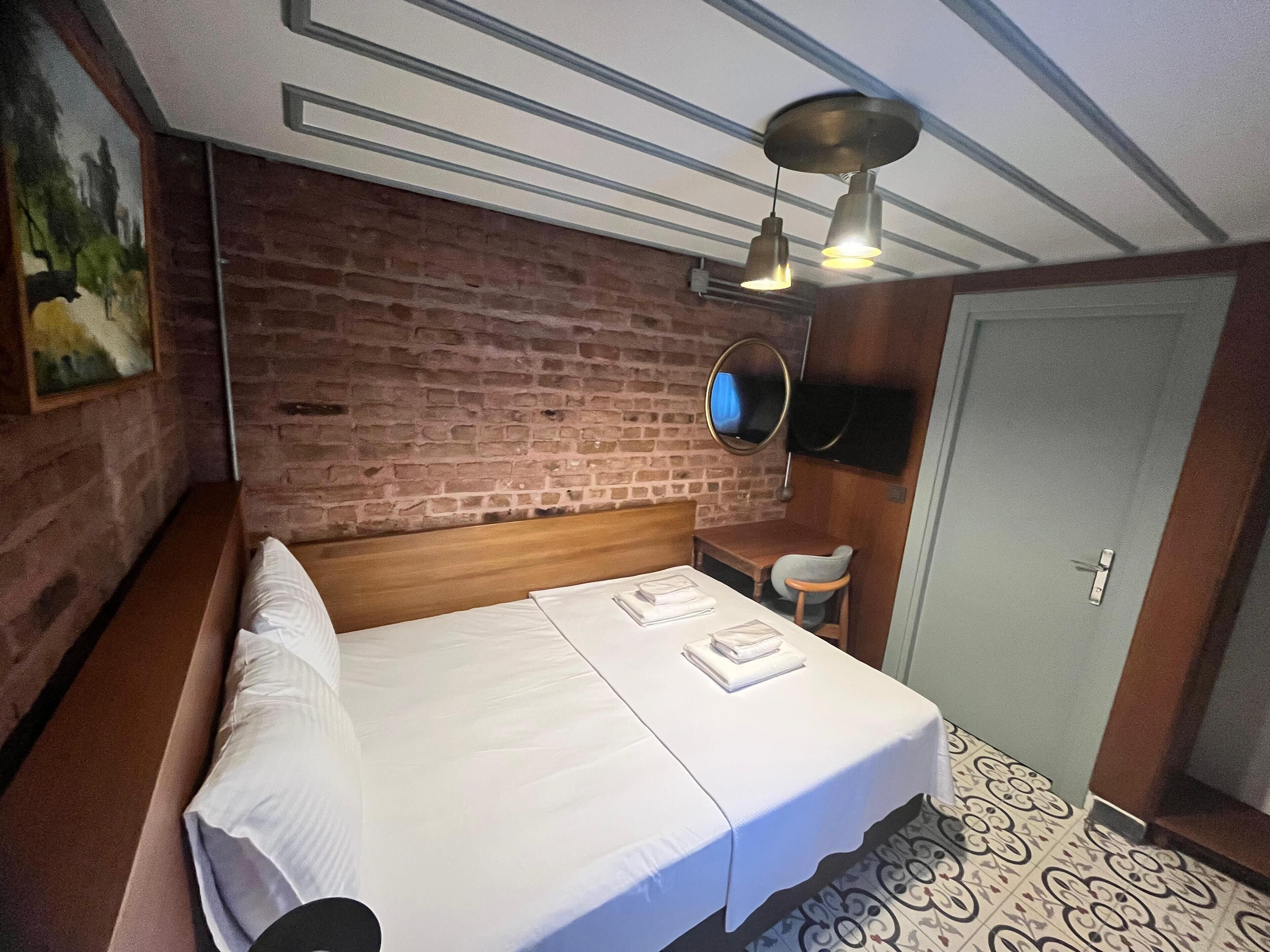 Habitación de hotel con pared de ladrillo, cama de madera, escritorio pequeño, una silla, televisor montado en la pared y una puerta.