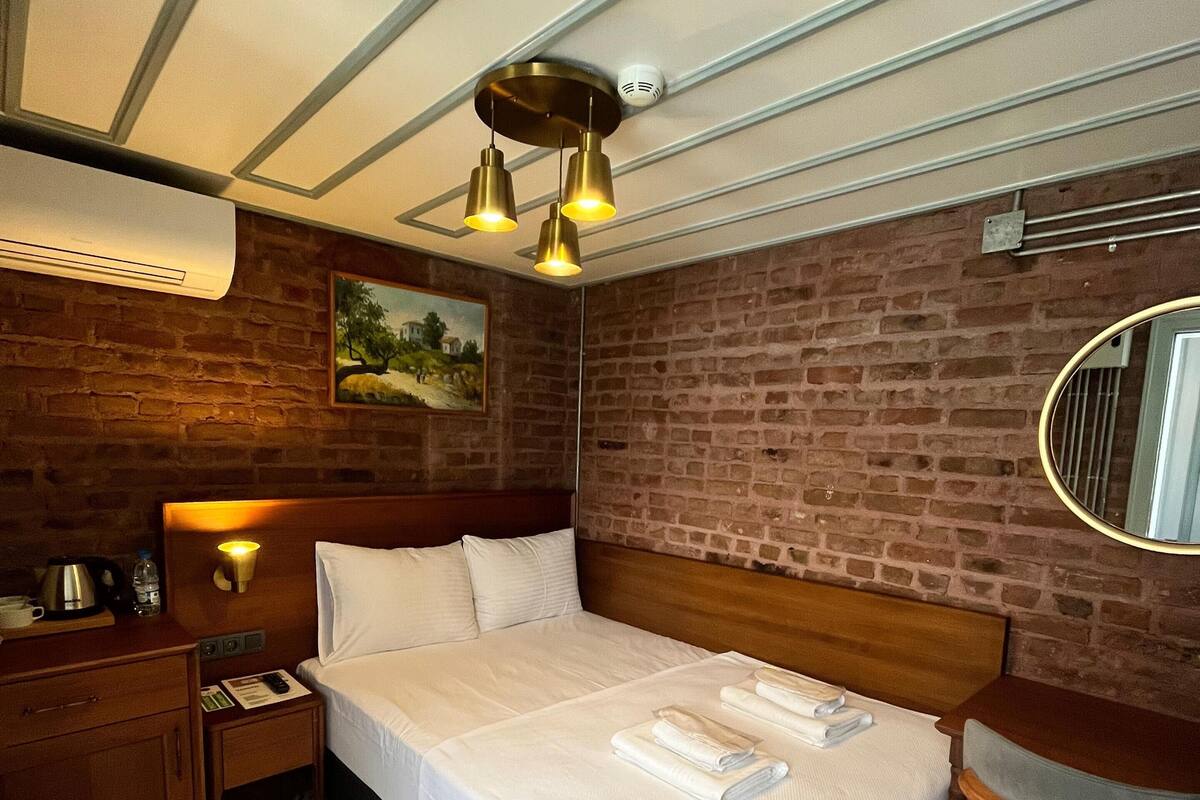 A hotel room with a brick wall, a bed with white linens, a wooden nightstand, a bedside table with a kettle, a wall-mounted light, and a patterned floor.