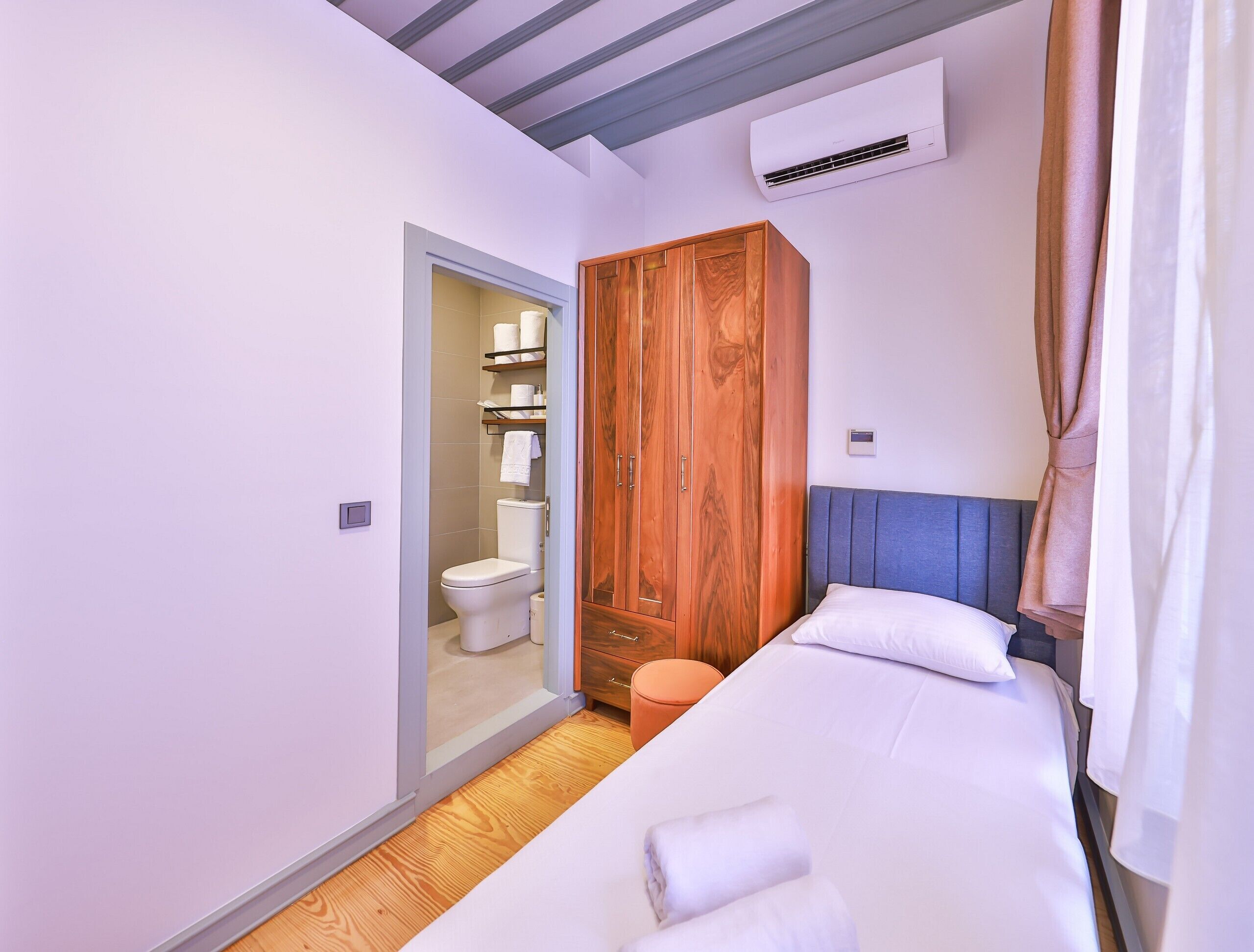 A bedroom with a wooden wardrobe, a bed with white linens, and a bathroom with a toilet and shelves.