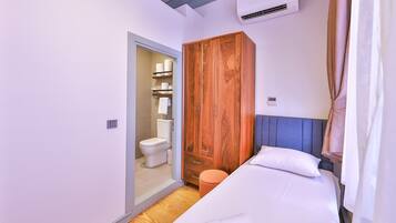 A bedroom with a wooden wardrobe, a bed with white linens, and a bathroom with a toilet and shelves.
