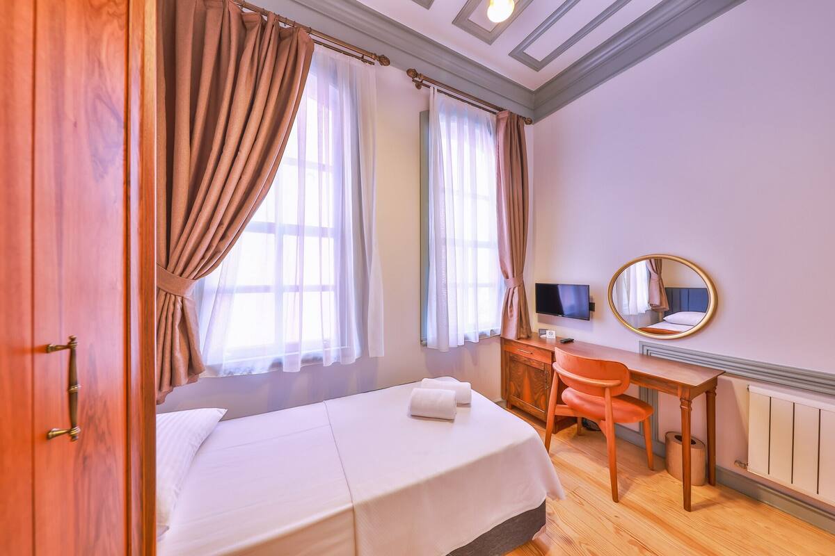 A bedroom with a bed, a desk, a chair, a mirror, and a window with curtains.