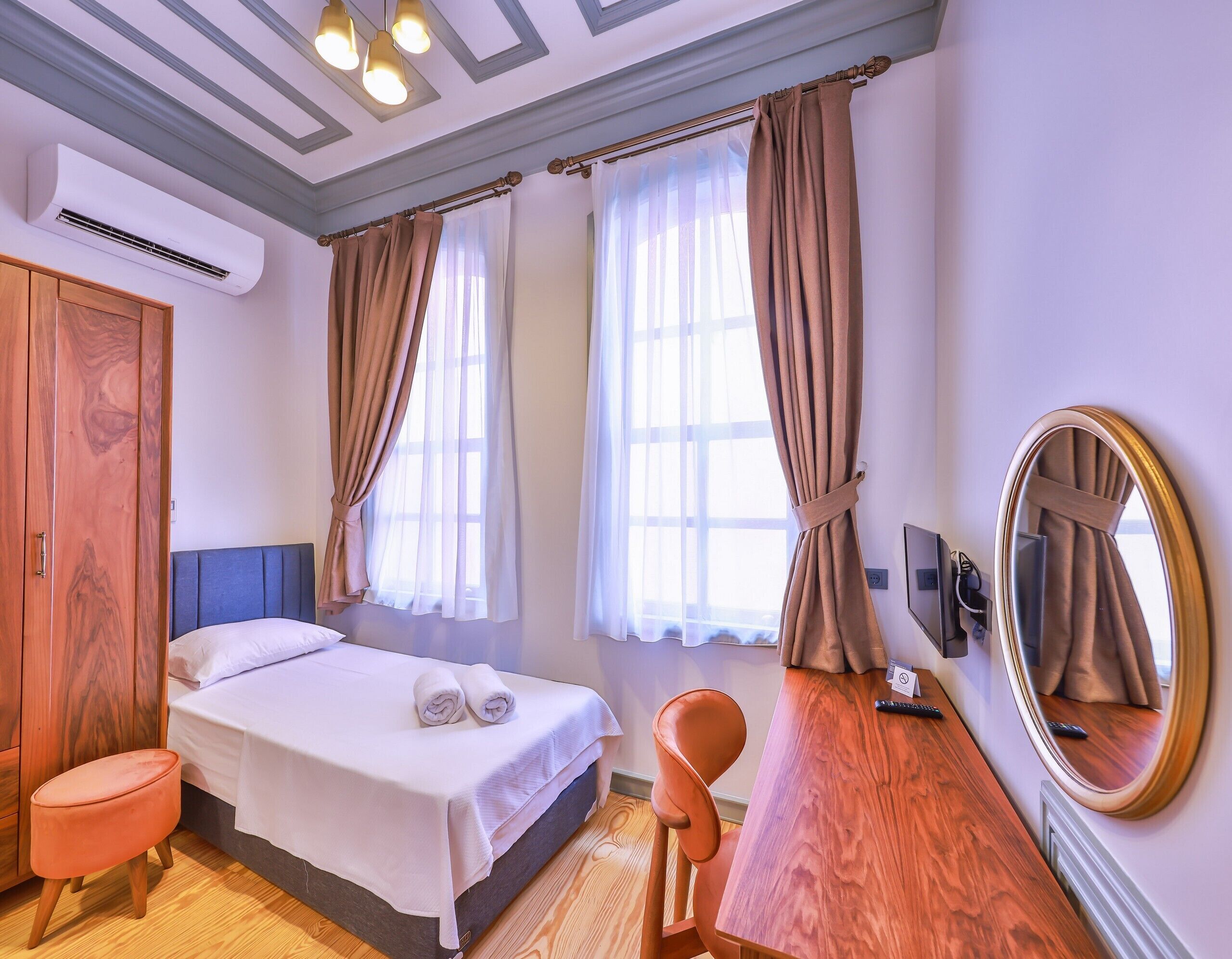 A hotel room with a bed, a wooden desk, a chair, a mirror, and a window with curtains.