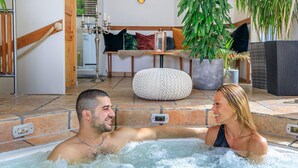 Un bain à remous avec un sol en pierre, un escalier en bois et un espace détente agrémenté de coussins et de plantes.
