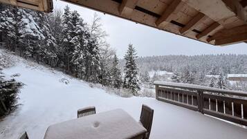 Une terrasse en bois avec une table et des chaises, offrant une vue sur un paysage enneigé, avec des arbres et un panorama lointain.