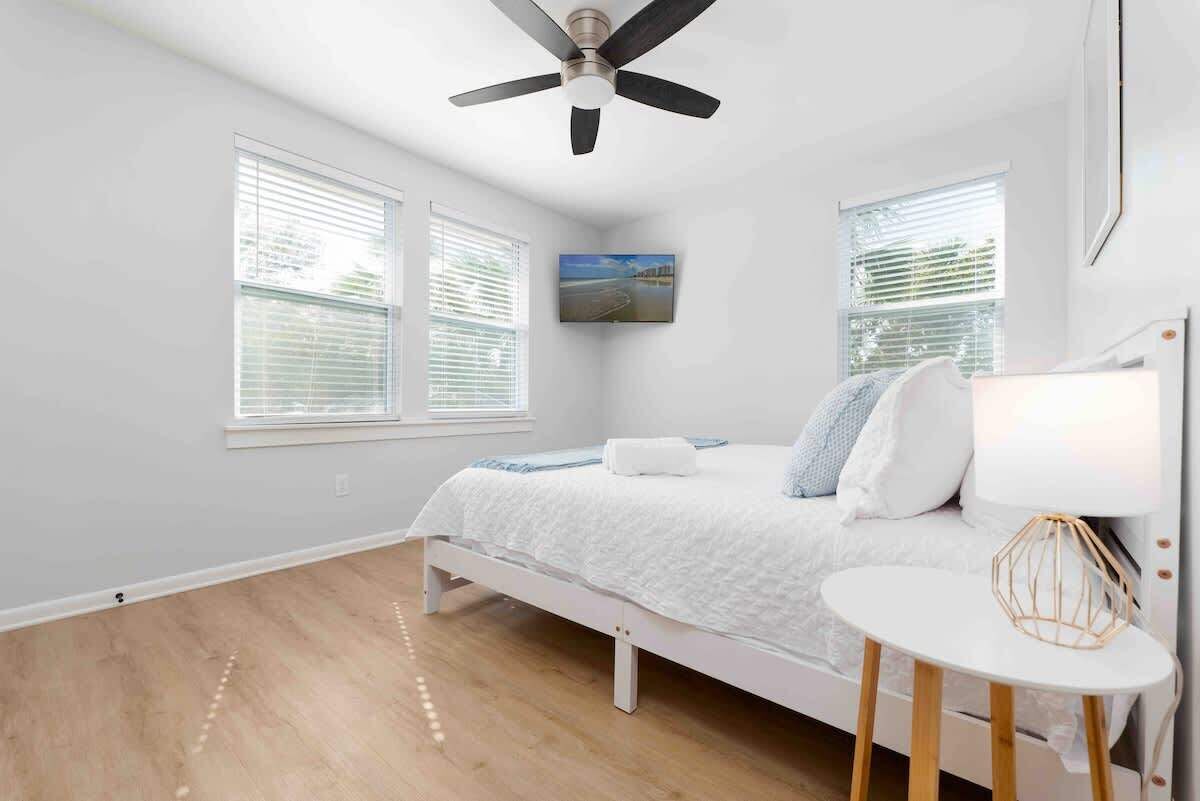 A bedroom with a bed, a ceiling fan, a TV mounted on the wall, and a bedside table with a lamp.