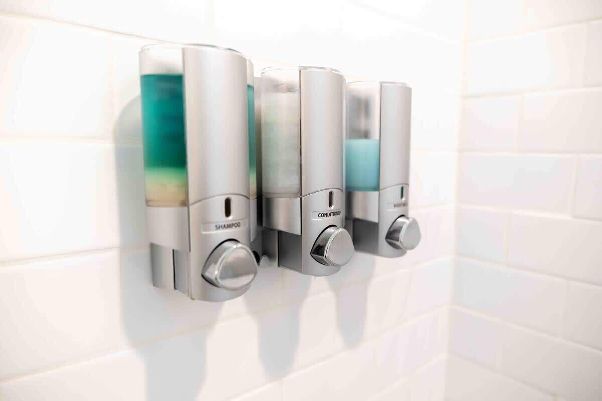 Un dispenser montato a parete con tre scomparti etichettati 'shampoo', 'balsamo' e 'detergente corpo'.