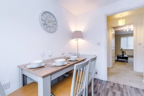 A dining area with a wooden table set for four, a wall clock, and a floor lamp. - Lovely three bed home Manchester (Bury)